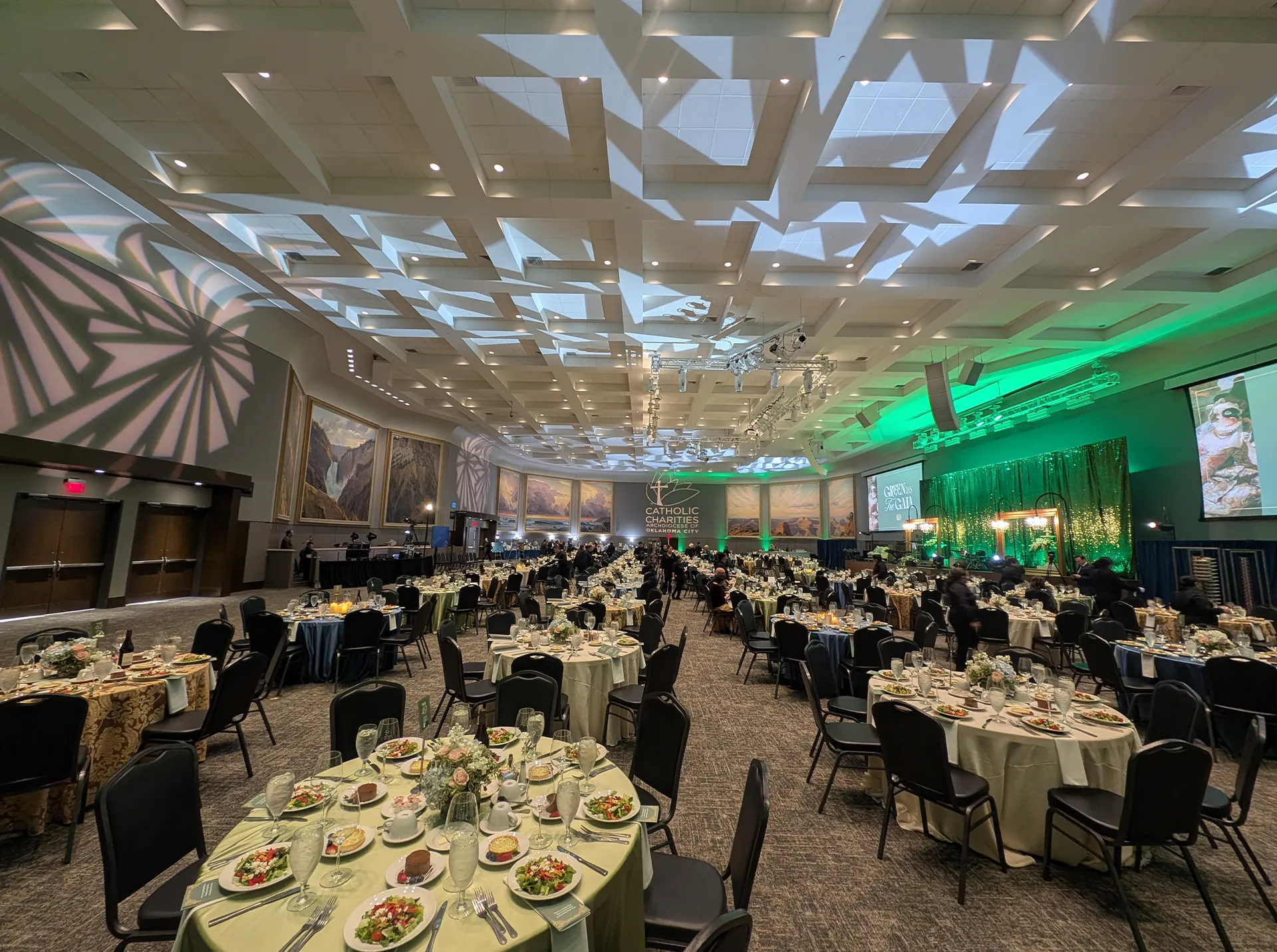 Ballroom set for the Green Tie Gala with green-lit stage, dinner tables, and Catholic Charities signage
